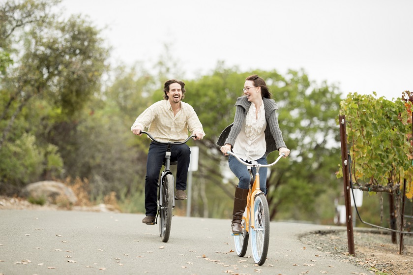 A smiling couple ride bikes past a vineyard on a cloudy day in Napa.